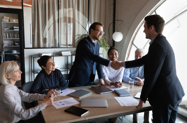 Five business professionals gathered around a conference table, two men in suits shaking hands while three others, two women and a man, observe and smile. Their discussion highlighted employer benefits, specifically the implementation of an Employment Assistance Program to provide mental health supports.