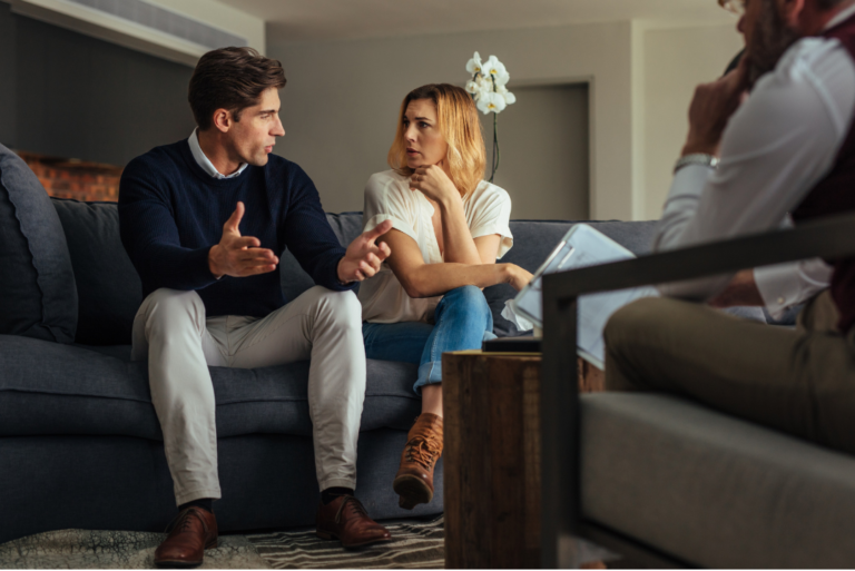 A man and a woman sit on a couch in a serious discussion about divorce counseling, with a third person observing them in the foreground.