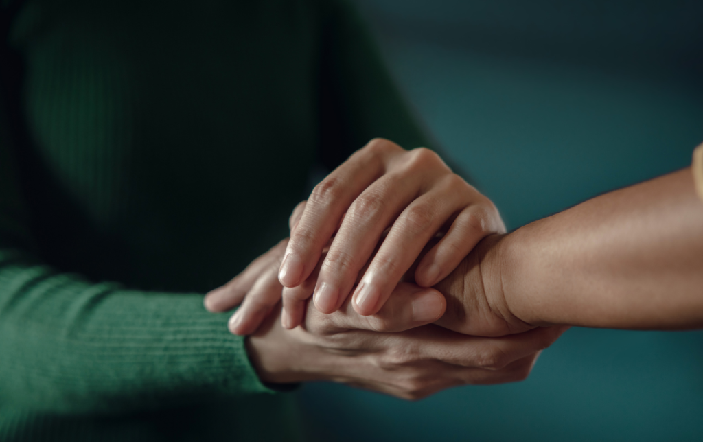 Close-up of two people holding hands. One person in a green sweater gently clasps the wrist of another person in a yellow shirt, evoking a moment of support often seen in divorce counseling. The background is blurred.