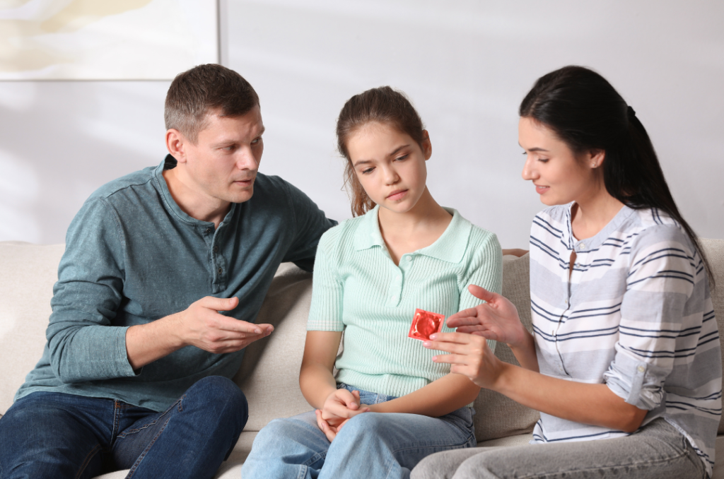 A man, woman, and teenage girl are seated on a couch. The woman is holding a red condom wrapper and explaining something to the girl as part of divorce counseling, while the man gestures with his hand.