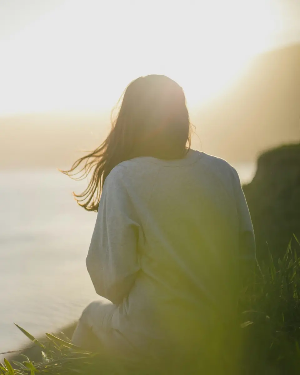 Woman sitting beside calm water at sunset, viewed from behind